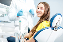 Portrait Of A Woman With Toothy Smile Sitting At The Dental Chair With Doctor On The Background At The Dental Office