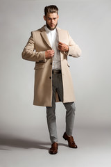 Elegant bearded man in a suit zips a beige coat, standing on a gray background