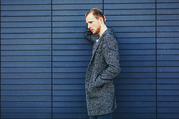 Wall Mural - Young stylish handsome man in coat posing next to a blue wall. copy space