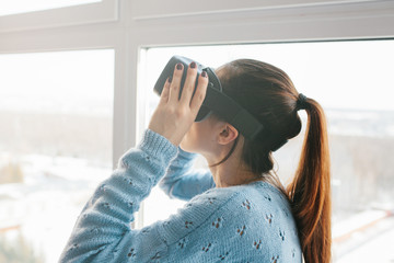 Beautiful young woman standing near a window in the smart home and looking into virtual reality goggles. Future technology concept. Modern imaging technology.