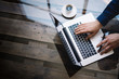 © SFIO CRACHO - Man working on laptop at sunny office.Male hand typing on keyboard.Modern notebook, cup of black coffee on the wooden table.Reflections on glass surface.Vertical.Top view.