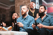 © mikhail_kayl - A company of four men with a beard sitting on the couch at home with beer and chips, two of them with joysticks in hand playing computer video games, the other carefully watching the game.