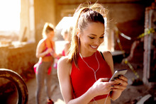 Young Attractive Fitness Model Listening Music On Smart Phone Charching Positive Energy Before Workout Outside. Sun Is Shining And Friends Are Behind Preparing For Training.