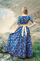 Beautiful Attractive Blond Caucasian Girl Model Stands Mountains and Sea Background in Long Blue Dress with Gold Yellow Pattern