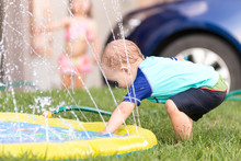 Child, Boy Or Kid Plays With Water Sprinkler Toy Outdoors During Summer Or Spring To Cool Off In Hot Weather