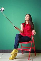 Wall Mural - Beautiful young woman taking selfie while sitting on chair near color wall