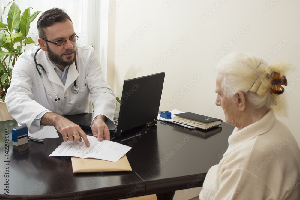 Lekarz geriatra przyjmuje pacjenta w gabinecie. Stara kobieta u lekarza geriatry. Lekarz tłumaczy pacjentce zapis w dokumentach. - obrazy, fototapety, plakaty 
