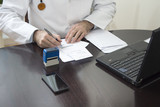 Fototapeta  - Lekarz w białym kitlu siedzi przy stole i przystawia pieczątkę do dokumentu. Ręce lekarza przystawiającego pieczątkę na recepcie. Lekarz wypisuje receptę przy biurku.