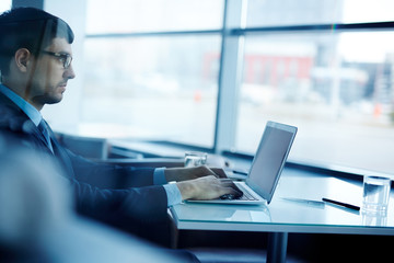 Profile view of concentrated young businessman in eyeglasses sitting at office desk and writing response letter to his partner on modern laptop, view through glass wall