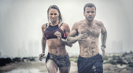 Muscular male and female athlete covered in mud running down a rough terrain with a desert background in an extreme sport race 