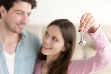 Smiling woman showing keys of apartment, looking at her husband. Young defocused couple preparing to move in their own house, real estate purchasing, successful property investment, focus on keys