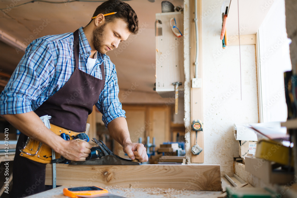 Profile view of confident young carpenter in apron planing board with jointer plane in spacious workshop