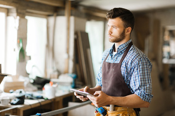 Wall Mural - Pensive young woodworker holding digital tablet in hands while looking out window of workshop, waist-up portrait