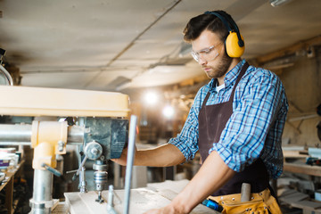 Wall Mural - Concentrated young woodworker in ear and eye protectors working with drill press in spacious workshop, waist-up portrait