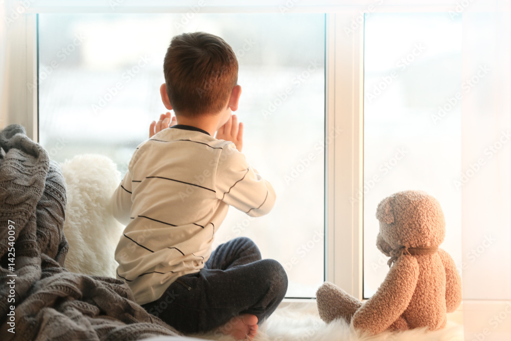 Cute little boy with teddy bear sitting on window sill at home