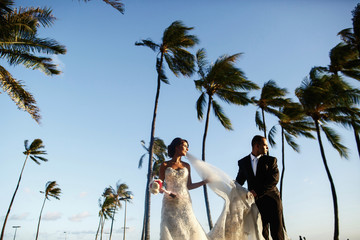 Look from below at wind blowing bride's veil away while she stands with groom under green palms