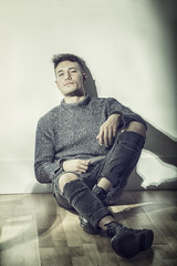 Wall Mural - Handsome white asian young man wearing grey sweater and ripped jeans, sitting on light background against wall in studio shot