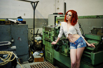 Wall Mural - Red haired girl wear on short denim shorts and white blouse posed at industrial machine at the factory.