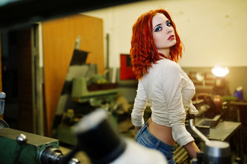 Wall Mural - Red haired girl wear on short denim shorts and white blouse posed at industrial machine at the factory.