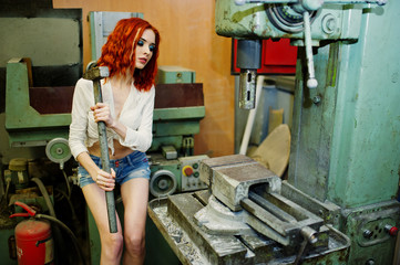 Wall Mural - Red haired girl wear on short denim shorts and white blouse with hammer at hands posed at industrial machine at the factory.