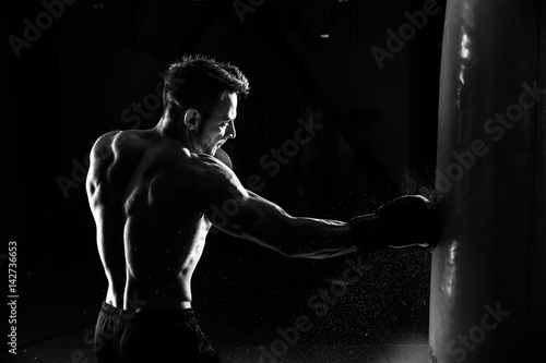 Leinwand Poster Young male boxer hitting punching bag on black background.
