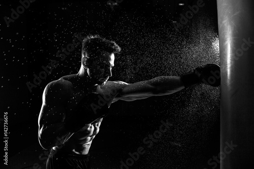 Leinwand Poster Young male boxer hitting punching bag on black background.