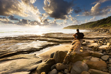 A Person Sits On A Beautiful Rocky Beach And Watches As The Sun Bursts Through The Clouds Over The Sea In This Beautiful Seascape.