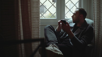 Stylish man sitting in armchair and looking at window. Young attractive male uses smartphone in living room.