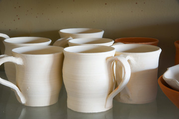 Ceramic or pottery greenware mugs on a glass shelf awaiting firing or bisque in contrasting light