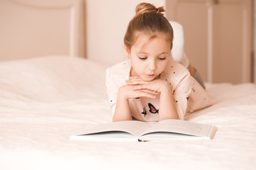 Sticker - Cute baby girl lying in bed reading book in room. Wearing pajamas at home. Childhood.