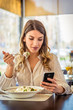 Young woman eating salad at restaurant and texting on smartphone