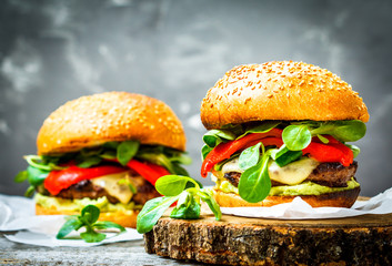 Canvas Print - Tasty grilled beef burger with cheese, guacamole and pickled peppers. Dark background, brown paper, wooden stand.