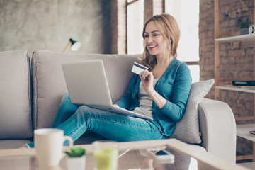 Concept of online shopping and payment.  Portrait of young smiling woman sitting on a sofa at home, holding credit card and using a computer for online shopping.