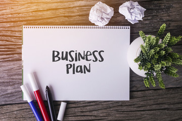 Wall Mural - Business Plan word with Notepad and green plant on wooden background.