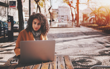 Attractive beautiful adult brunette freelancer girl with black curly hair sitting in street cafe and working on her laptop with copy space for young advertising text message or promotional content