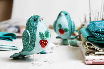 Closeup a handmade bird sewed from a cloth stands on a white wooden table surrounded by handicraft  materials. Shallow focus and blurred background.