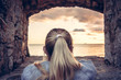 Thoughtful woman devoted into contemplation of beautiful sunset over sea through window of old castle with dramatic sky and perspective view. View from behind. Concept for forecasting and way forward