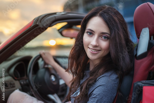 Pretty young brunette woman driving luxury red cabriolet car at the sunset Wallpaper Mural