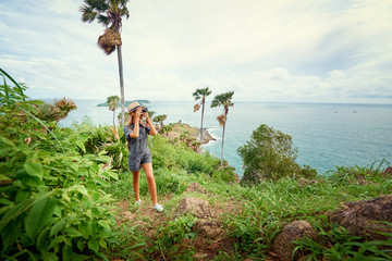 Wall Mural - Photography and travel. Young woman in hat with rucksack taking picture on her camera enjoying sea view.