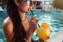 Beautiful Brunette Woman Wearing A Blue Bikini And Jeans Shorts, Enjoying The Pool, Summer Time.Sensual Brunette Beautiful Girl With Perfect Body Drinking Cocktail While Relaxing Near The Pool