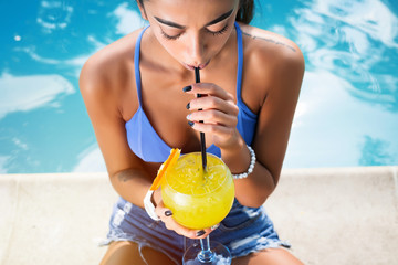 Wall Mural - Beautiful brunette woman wearing a blue bikini and jeans shorts, enjoying the pool, summer time.Sensual brunette beautiful girl with perfect body drinking cocktail while relaxing near the pool