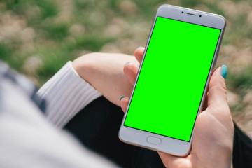 Wall Mural - Girl with smartphone in hands with mock up green screen of blank screen sits in park on open space, screen for content integration. Hands holding gadget on blurred backdrop, front view