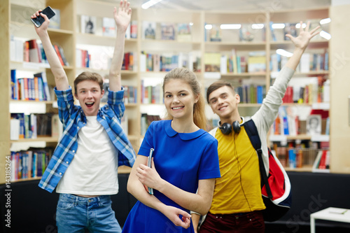 Group Of Three Hy Students Smiling Cheerfully And Raising Hands Group Of Three Hy Students Smiling Cheerfully And Raising Hands In Library Modern College