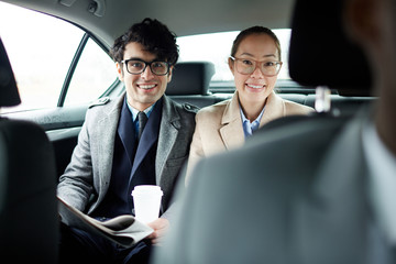 Portrait of two successful business people riding on backseat of taxi: smiling cheerfully and looking at camera on the way to work