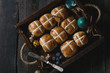 Hot cross buns in wooden tray served with butter, knife, blueberries, easter eggs, birch branch over old texture wood background. Top view, space. Easter baking.