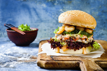 Wall Mural - Asian style salmon burger with grilled shrimps, seaweed, lettuce and spicy sriracha mayo sauce served on pieces of brown paper on a rustic wooden board. Bright blue background with copyspace