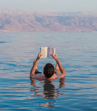Young Man Reads A Book Floating In The Waters Of The Dead Sea In Israel
