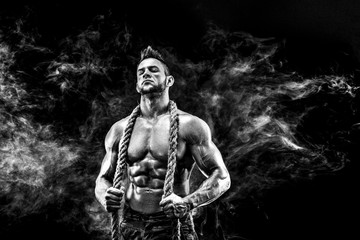 strong athletic man with naked body in military pants and rope on neck dark with smoke background