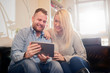 © chika_milan - Happy couple smiling and sitting on sofa in living room. They using tablet
