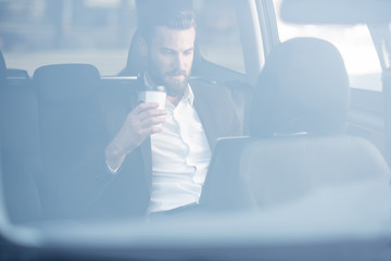 Handsome businessman sitting with coffee to go on the backseat of the car. View through the window